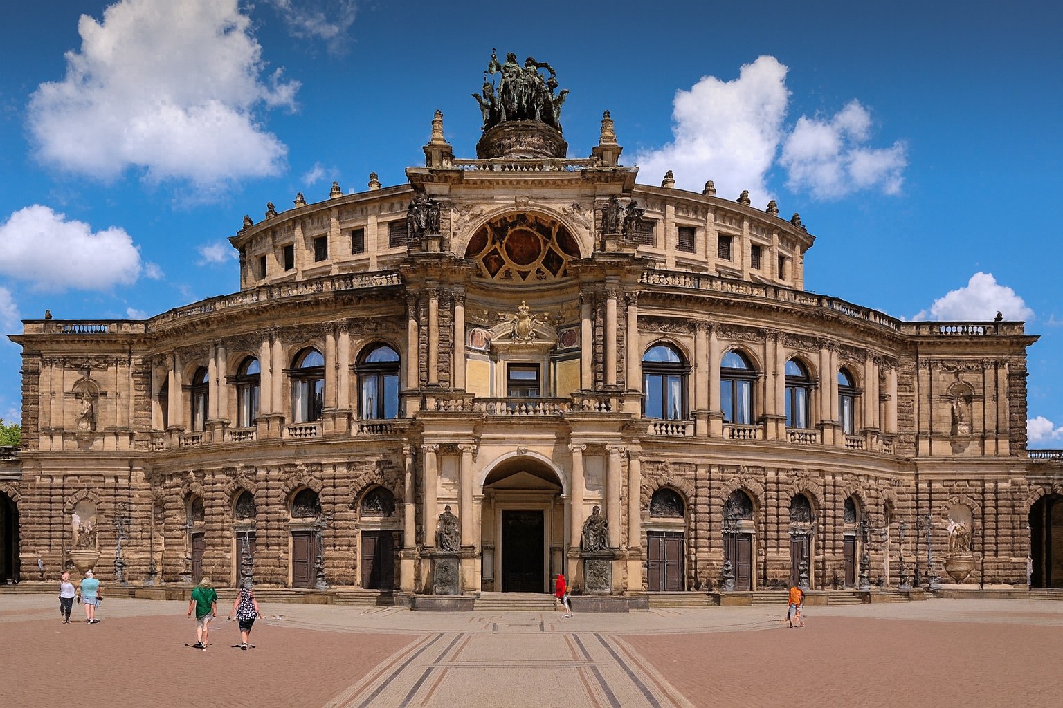 Semperoper in Dresden in Deutschland mit reich verzierter Fassade im Stil der Neorenaissance, bei bestem Sonnenschein mit malerischen Wolken, belebter Vorplatz mit Spaziergängern.