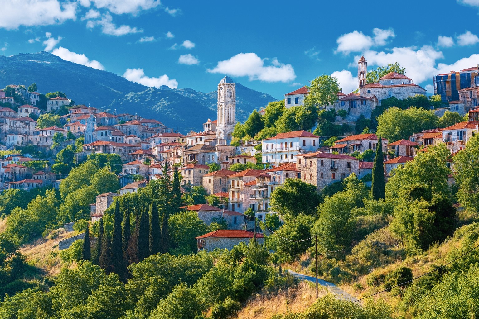 Panorama-Bild von Edessa mit traditionellen Steinhäusern und roten Ziegeldächern, eingebettet in eine grüne Hügellandschaft, im Hintergrund die Berge unter blauem Himmel mit weißen Wolken.