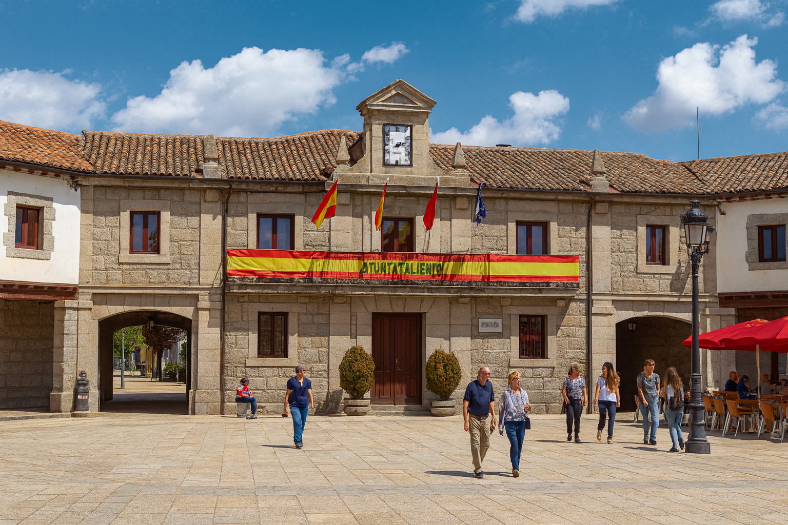 Plaza Mayor in Guadarrama mit der Stadthalle, Spaziergängern und Cafétischen im Sonnenschein unter malerischen weiß-grauen Wolken.