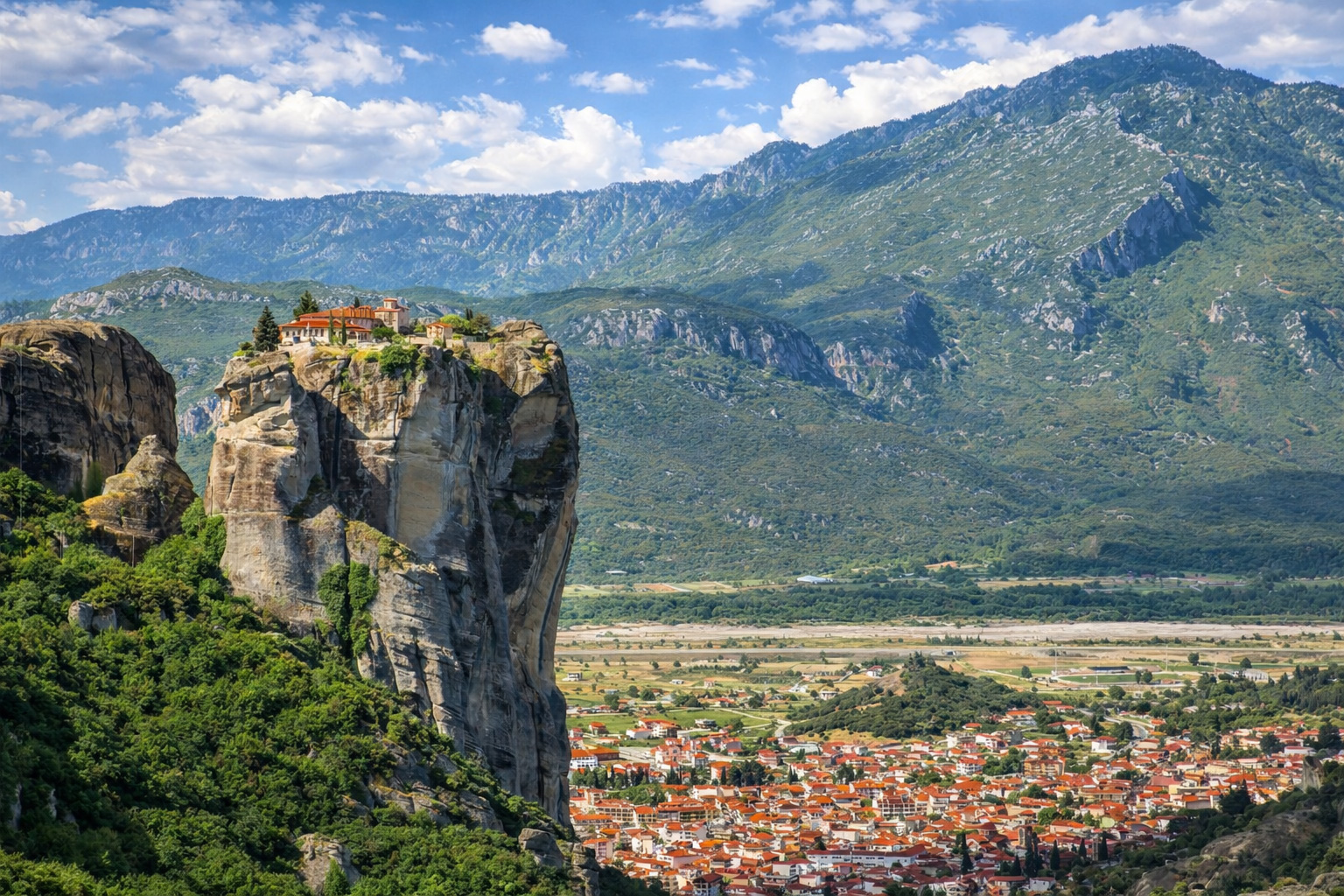 Panoramaaufnahme des Meteora-Klosters bei Kalambaka in Griechenland mit markantem Felsen, Klosteranlage und weißen Wolken über den Bergen.