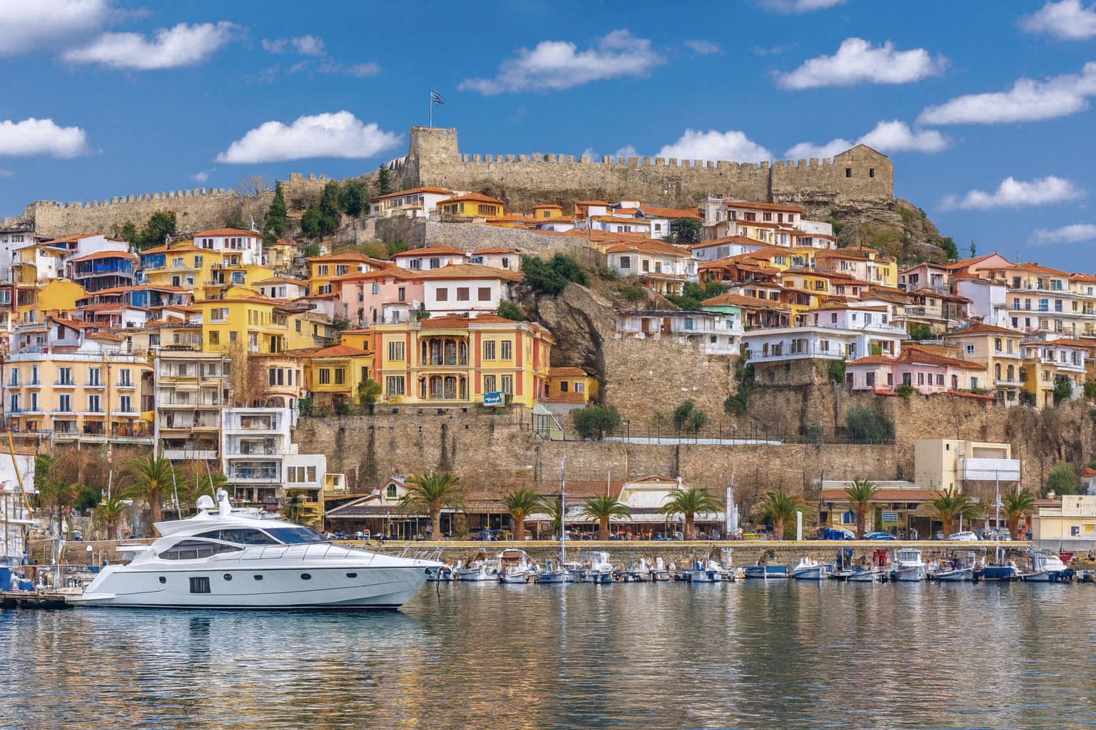 Panorama von Kavala mit bunten Häusern am Hang, Festungsmauern und Hafen mit moderner Motoryacht unter blauem Himmel mit weißen Wolken.