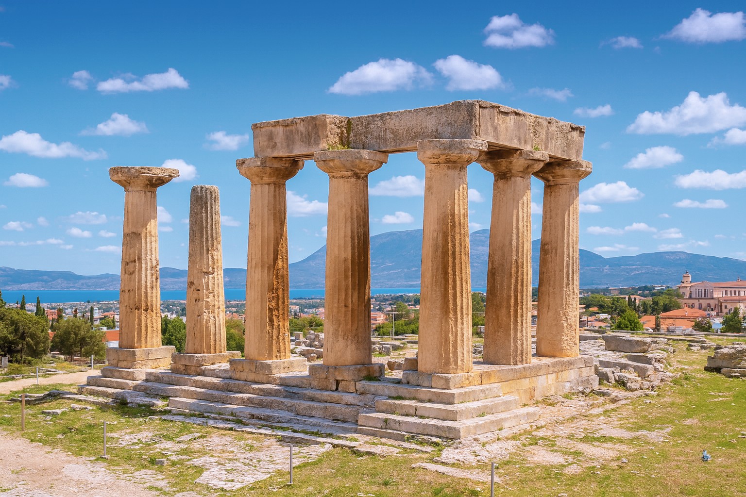 Panorama des antiken Apollon-Tempels in Korinth mit dorischen Säulen, Ausgrabungsstätte und Blick auf Meer, Stadt und Berge unter blauem Himmel mit weißen Wolken.