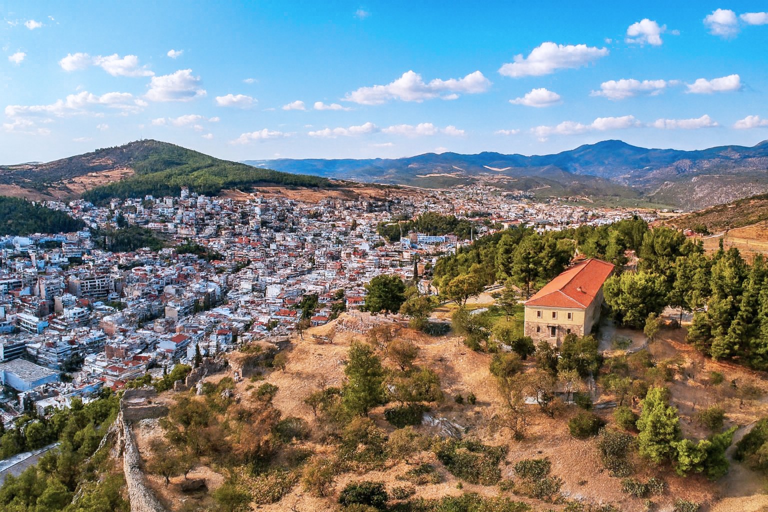 Panoramaaufnahme der Stadt Lamia mit der Akrolamia-Burg auf einem Hügel, umgeben von grünen Bäumen und Bergen, unter blauem Himmel mit weißen Wolken.