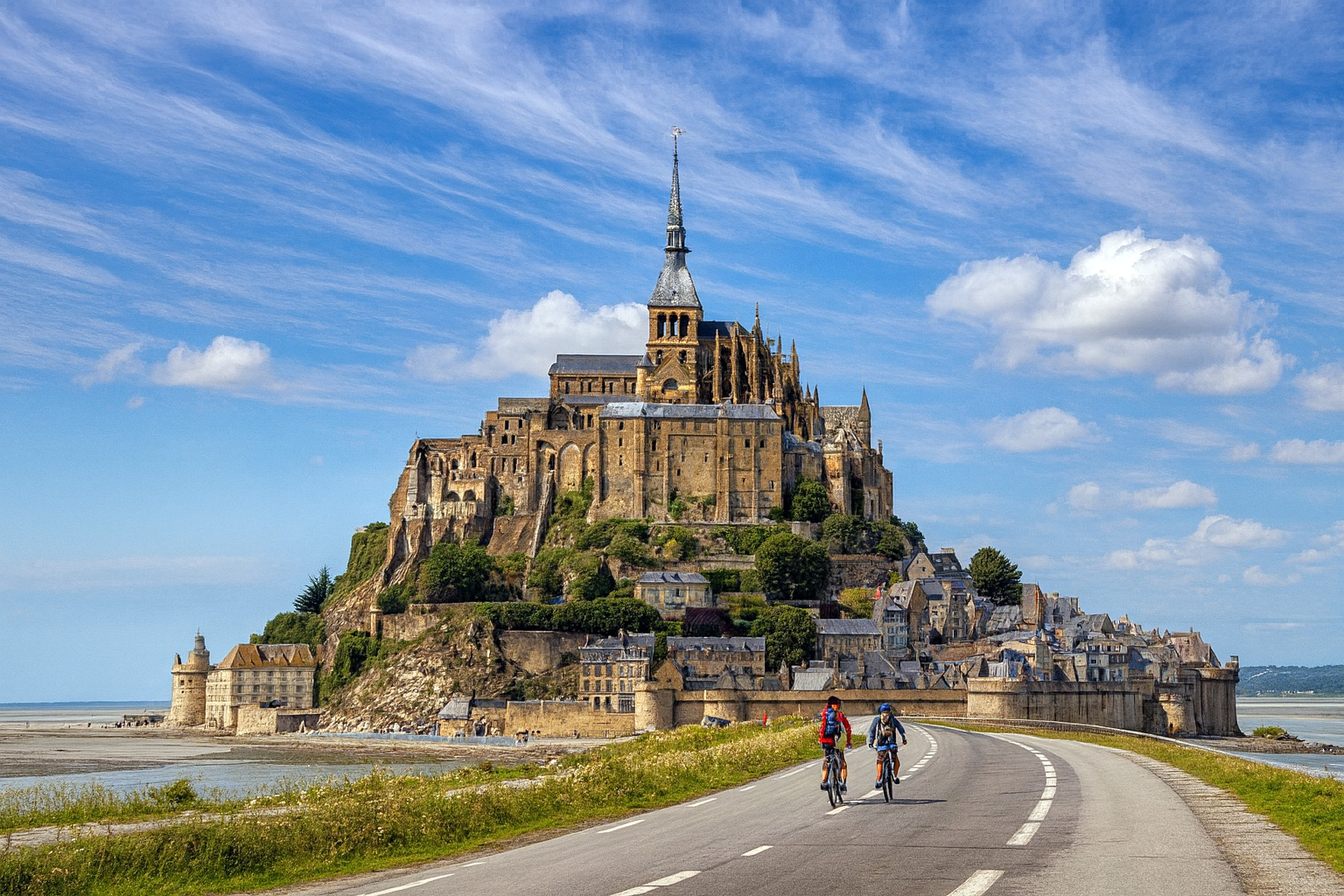 Panorama von Le Mont-Saint-Michel mit zwei Fahrradfahrern auf der Straße im Vordergrund unter einem Himmel mit malerischen Cumuluswolken und Sonnenlicht.