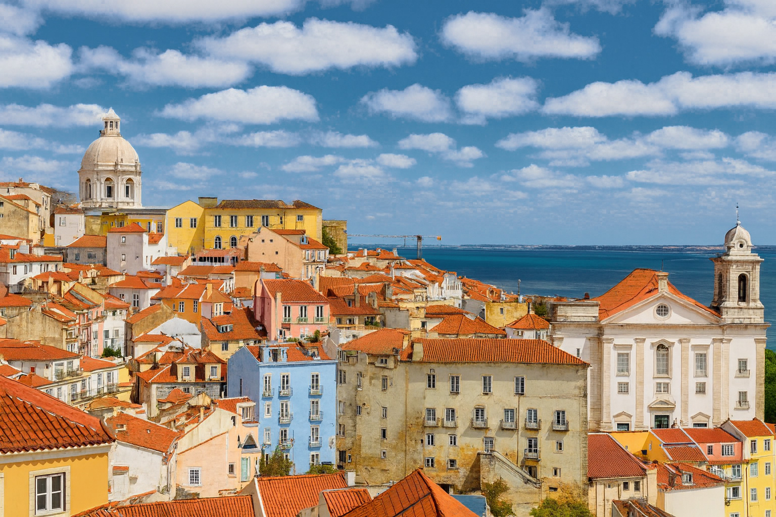 Panoramaaufnahme von Lissabon mit Blick auf die Igreja de Santa Engrácia, bunten Häusern mit roten Dächern und dem Meer im Hintergrund bei klarem Sonnenlicht und malerischen weiß-grauen Wolken.