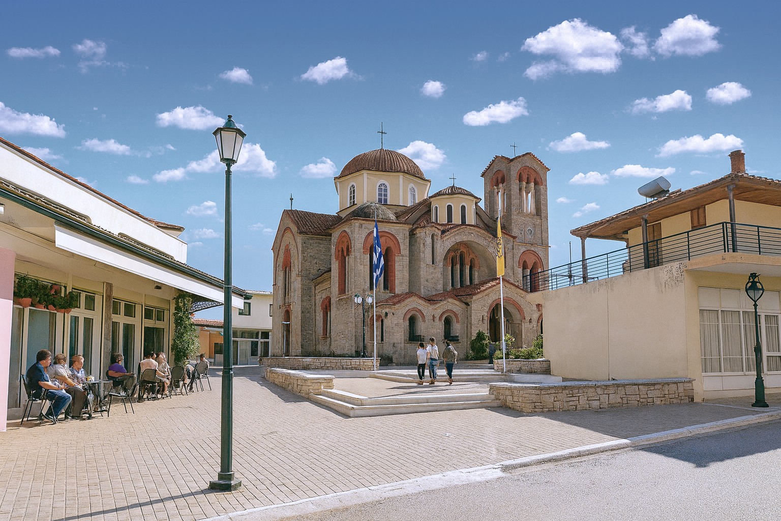 Platz in der Kleinstadt Livanates mit der St.-Georg-Kirche im Hintergrund, Straßencafé mit Gästen und blauem Himmel mit weißen Wolken.