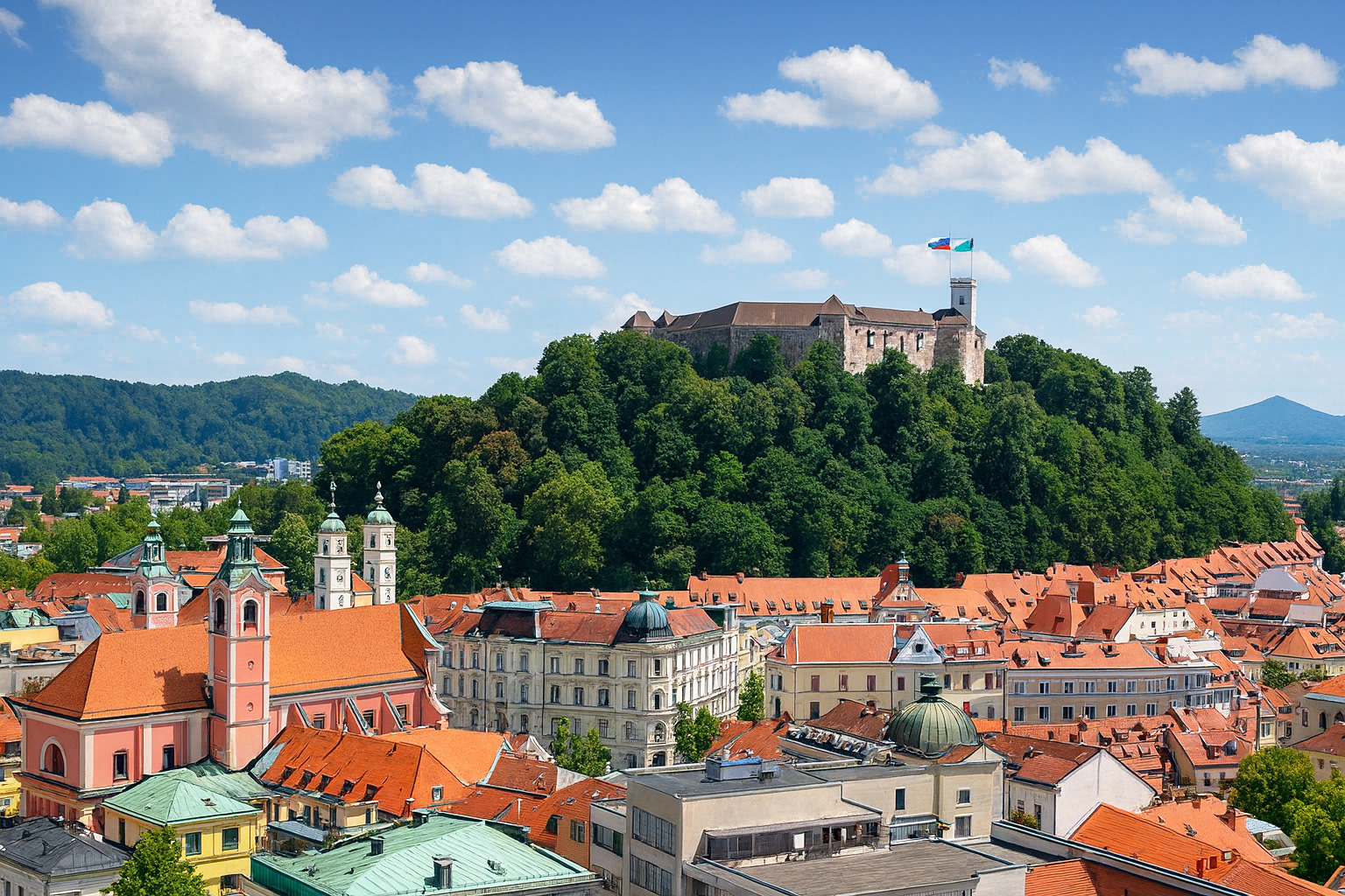 Blick über die Altstadt von Ljubljana mit der Burg auf dem Schlossberg und roten Ziegeldächern bei blauem Himmel mit weißen Wolken