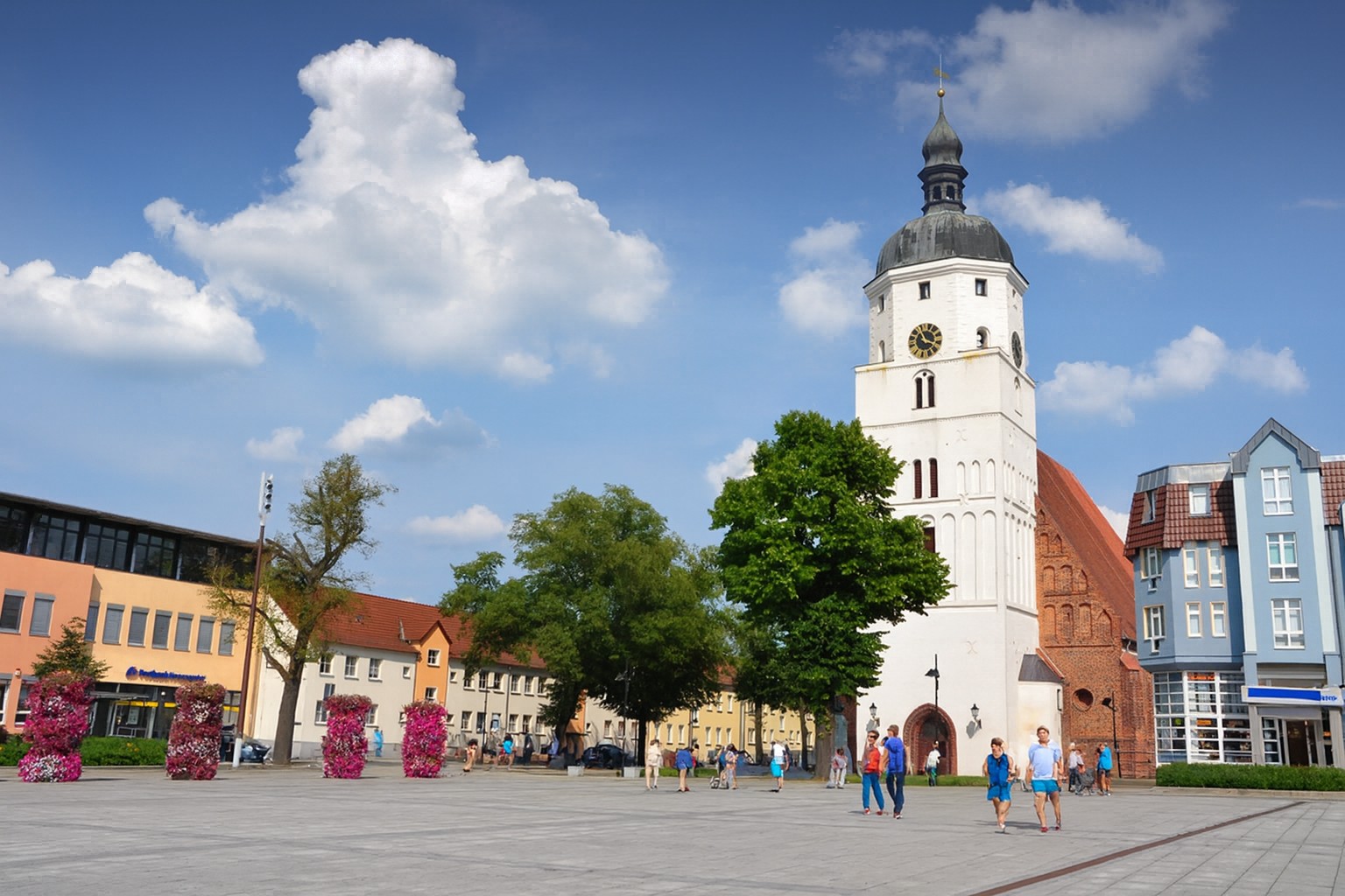 Marktplatz in Lübben in Deutschland mit der Kirche und ihrem markanten Turm im Hintergrund, bei bestem Sonnenschein mit malerischen Wolken, belebt mit Spaziergängern.