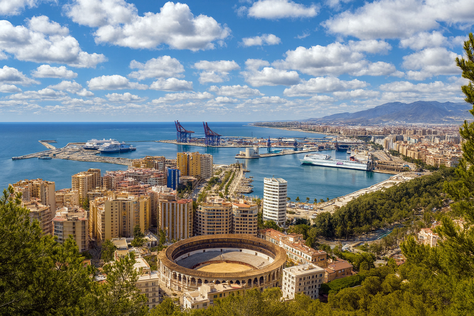 Panoramaaufnahme von Málaga mit der historischen Stierkampfarena, dem Hafen und dem Mittelmeer bei klarem Sonnenlicht und malerischen weiß-grauen Wolken am Himmel.