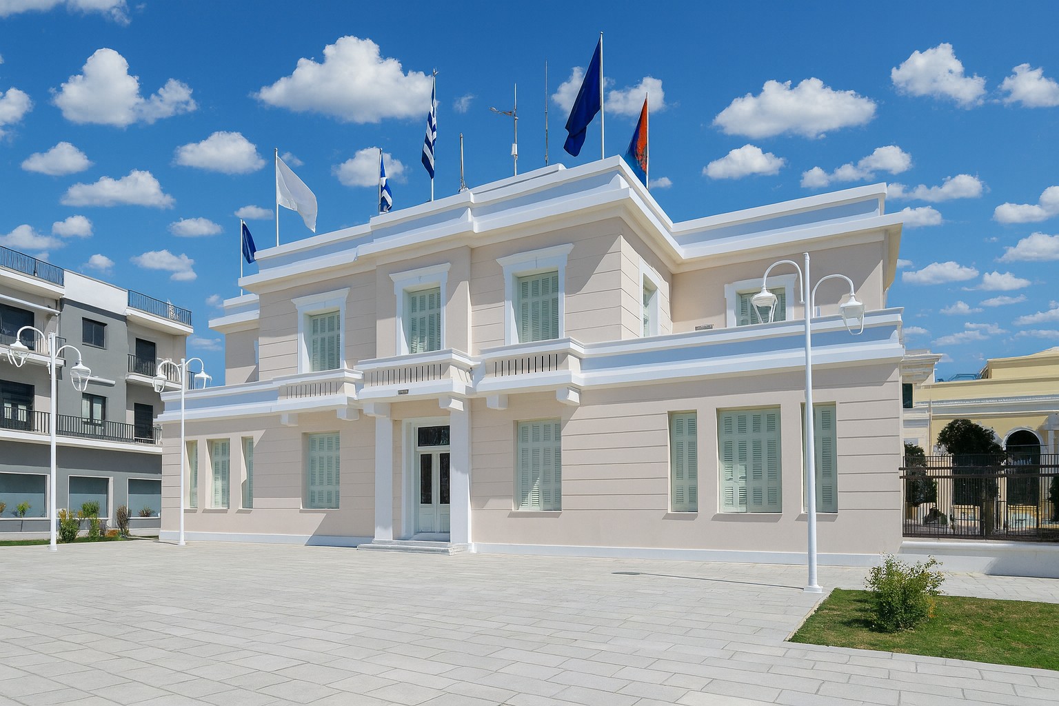 Museum für Geschichte und Kunst in Mesolongi mit neugestaltetem Vorplatz aus hellen Pflastersteinen, ohne Personen und Fahrräder, bei Tageslicht mit weißen Wolken am Himmel.