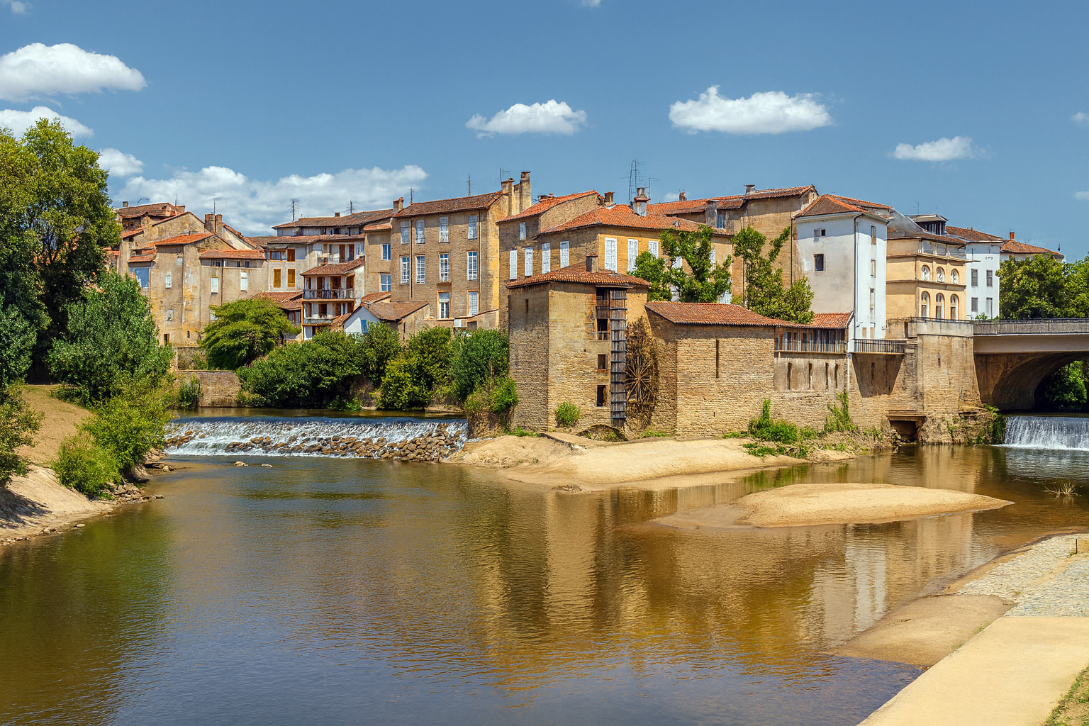Panorama der Altstadt von Mont-de-Marsan mit historischen Gebäuden am Fluss Midou und kleinen Wasserfällen unter blauem Himmel mit weißen Wolken.
