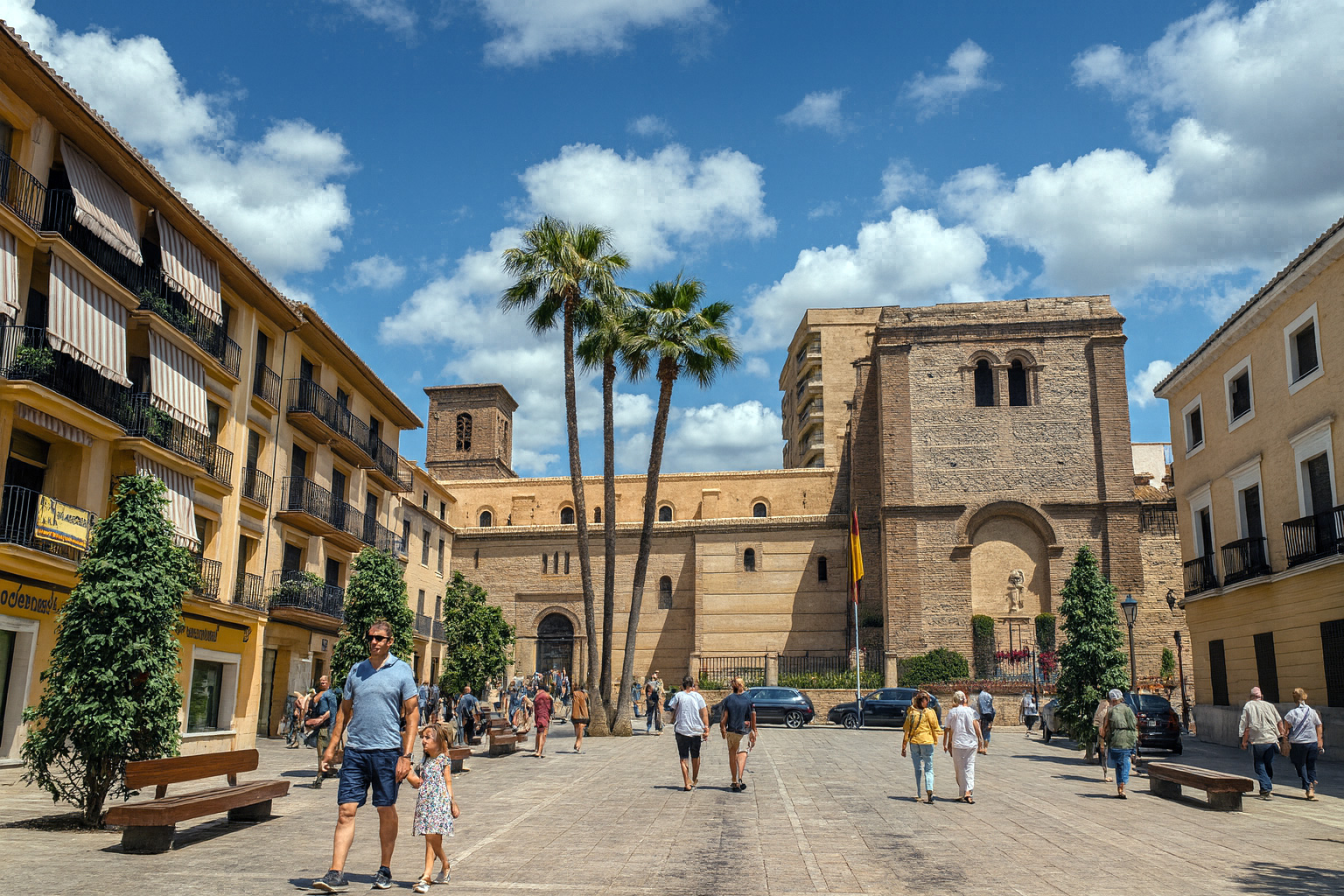 Plaza de España in Motril mit der historischen Kirche und Palmen bei klarem Sonnenlicht, einige Spaziergänger schlendern über den Platz unter malerischen weiß-grauen Wolken.