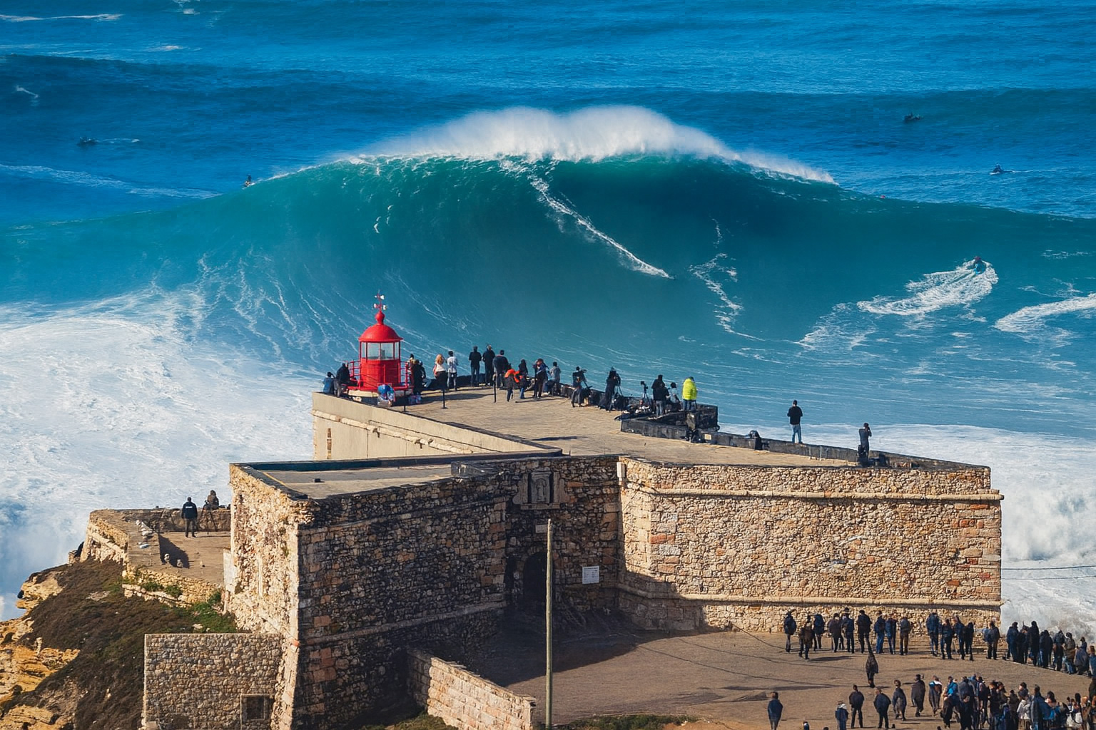 Aussichtspunkt Farol da Nazaré mit Zuschauern auf der Festung, während sich gewaltige Atlantikwellen brechen, bei klarem Himmel und strahlendem Sonnenlicht.