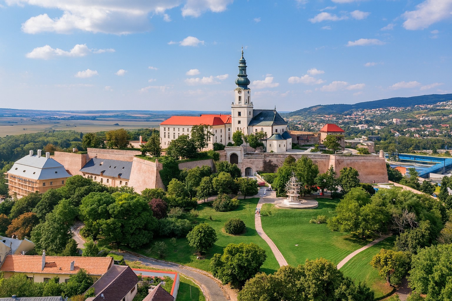 Panorama der Burg in Nitra mit Barockkirche und Festungsmauern, grüner Parkanlage und weißen Wolken am blauen Himmel in der Mittagssonne.