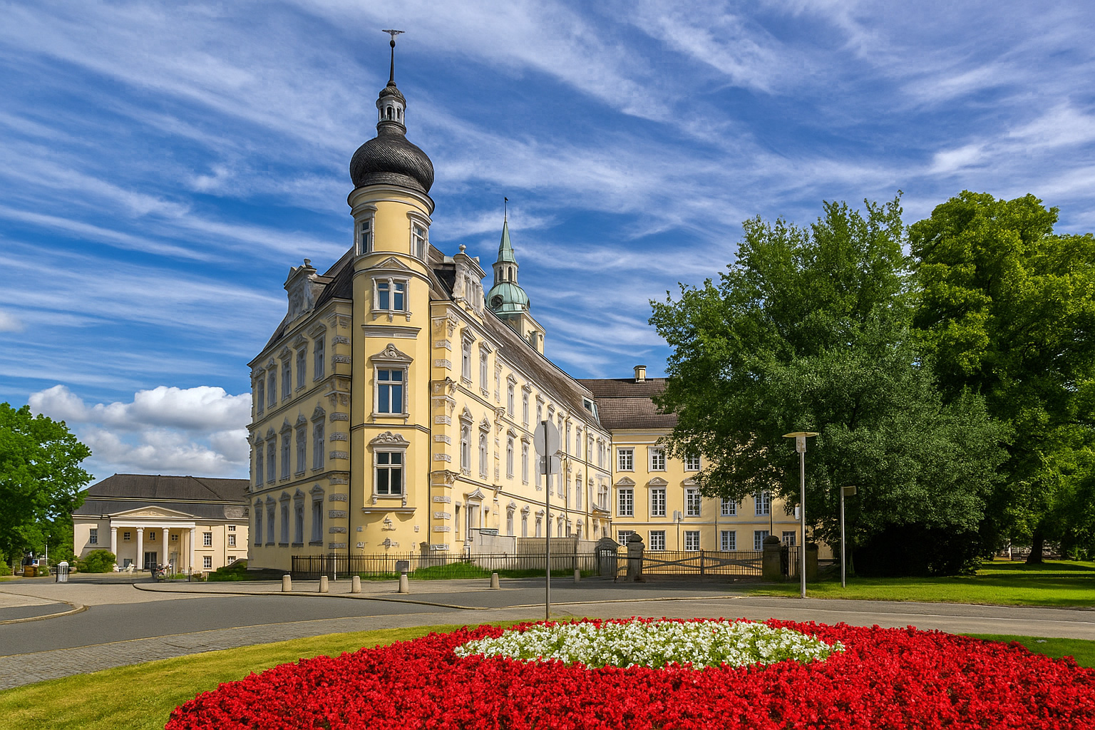 Blick auf das Oldenburger Schloss mit dem Schlossplatz und einem Blumenbeet im Vordergrund unter einem Himmel mit Zirrus- und Kumuluswolken.