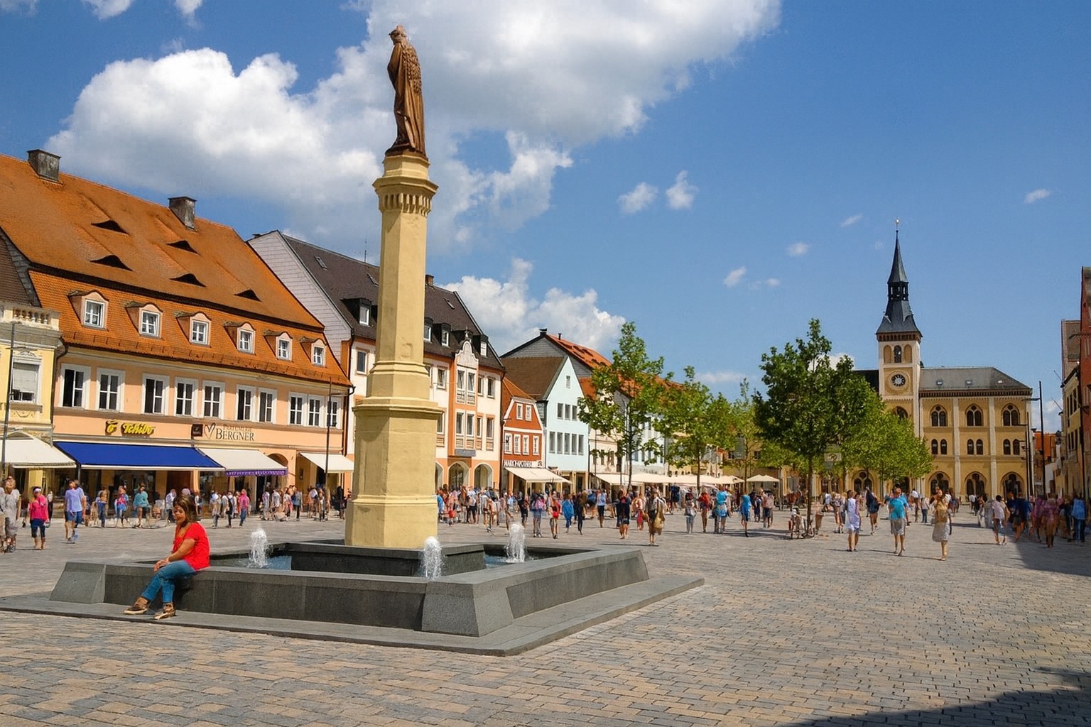 Marienbrunnen auf dem Hauptplatz in Pfaffenhofen an der Ilm mit Blick auf das historische Rathaus, belebt mit Spaziergängern.