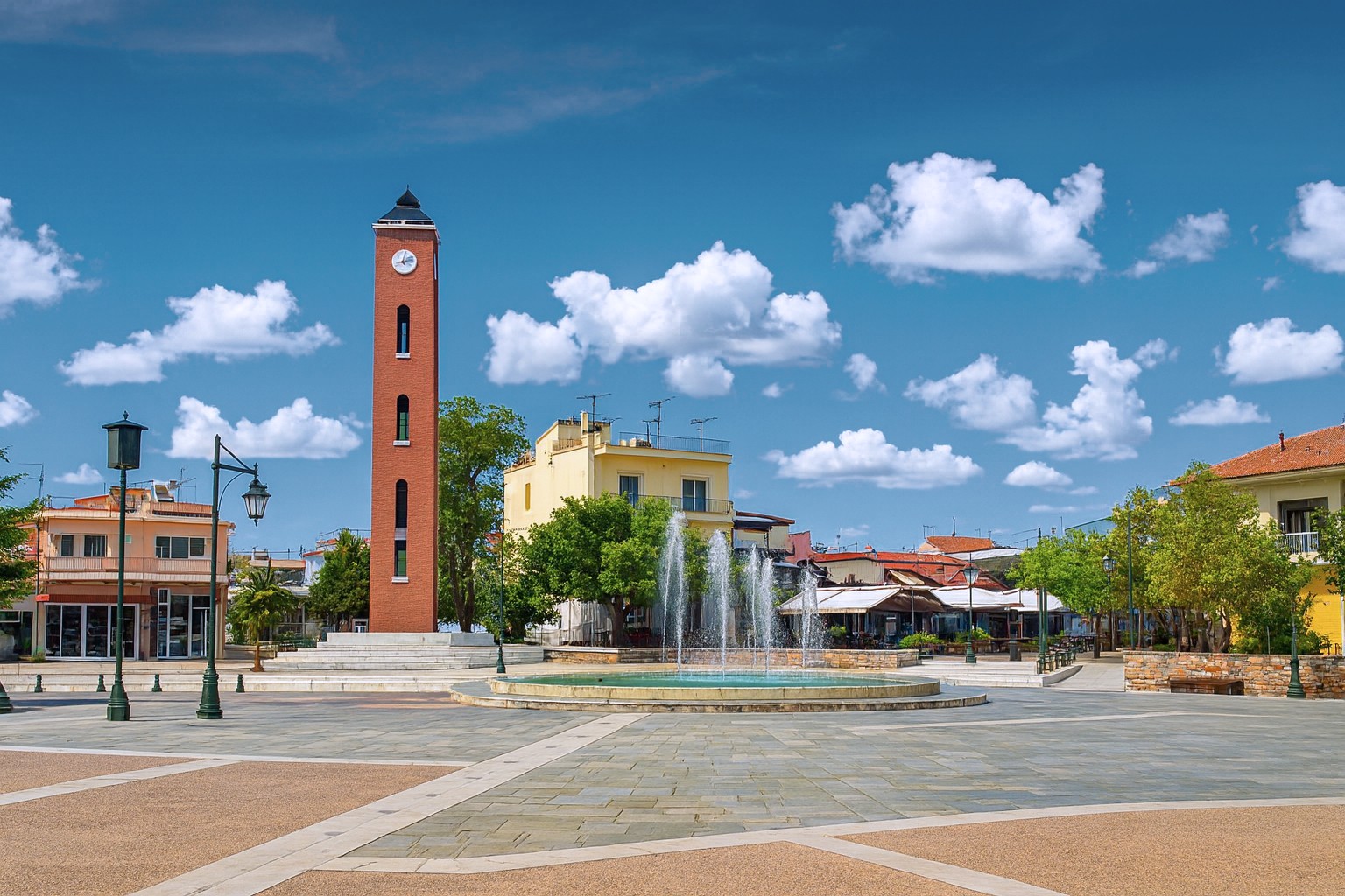 Hauptplatz in Polykastro mit rotem Uhrturm und Brunnen, bei bestem Tageslicht, umgeben von Straßencafés und Gebäuden, unter blauem Himmel mit weißen Wolken.