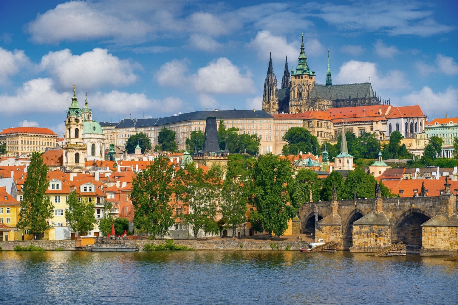 Panorama von Prag mit Burg, St. Veitsdom, St. Nikolauskirche und Karlsbrücke an der Moldau, bei strahlender Mittagssonne mit malerischen weißen Wolken.