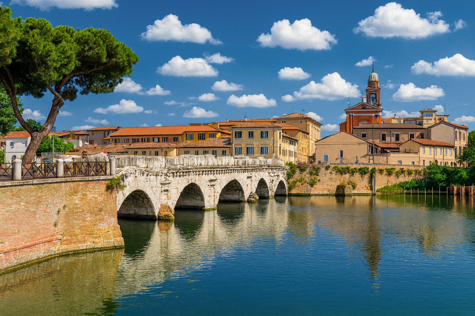 Tiberiusbrücke in Rimini mit historischen Gebäuden und der Kirche San Giovanni Battista im Hintergrund bei blauem Himmel mit weißen Wolken.