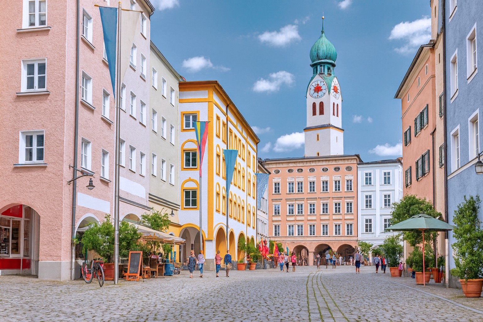 Altstadt von Rosenheim mit Blick auf den Max-Josefs-Platz, bunten Bürgerhäusern und dem Kirchturm von St. Nikolaus im Hintergrund, belebt mit Spaziergängern.
