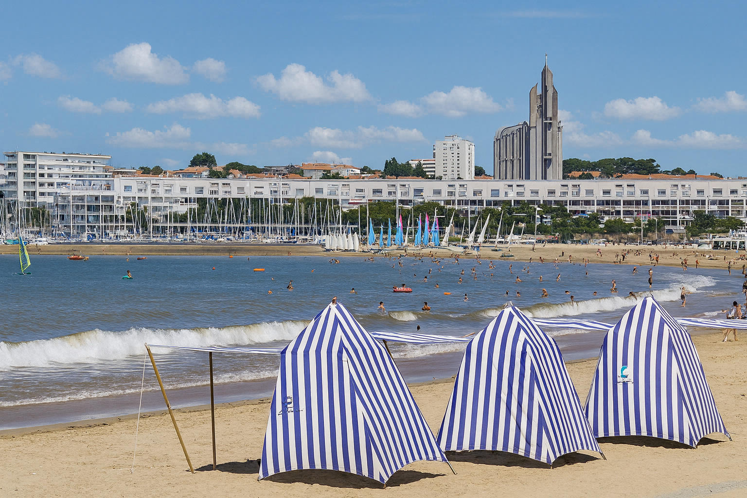 Sommerlicher Strand in Royan mit blau-weiß gestreiften Strandzelten, Badegästen im Meer und der modernen Kirche Église Notre-Dame de Royan im Hintergrund.