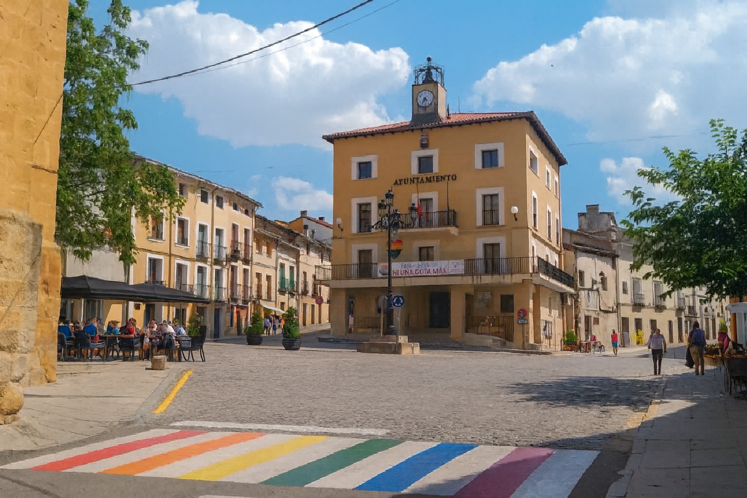 Plaza de la Constitución in Sacedón mit dem Rathaus und einem Straßencafé unter Sonnenschirmen bei sonnigem Wetter und malerischen weiß-grauen Wolken.
