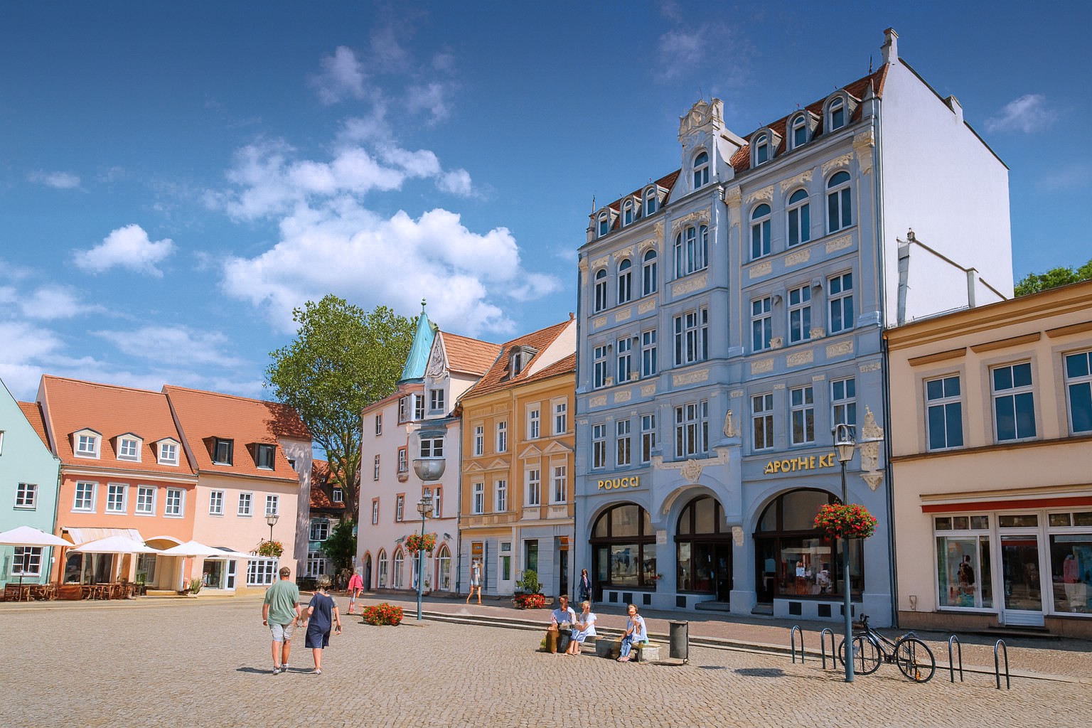 Marktplatz in Senftenberg in Deutschland mit farbenfrohen Fassaden und der Adler Apotheke, bei bestem Sonnenschein mit malerischen Wolken, belebt mit wenigen Spaziergängern.