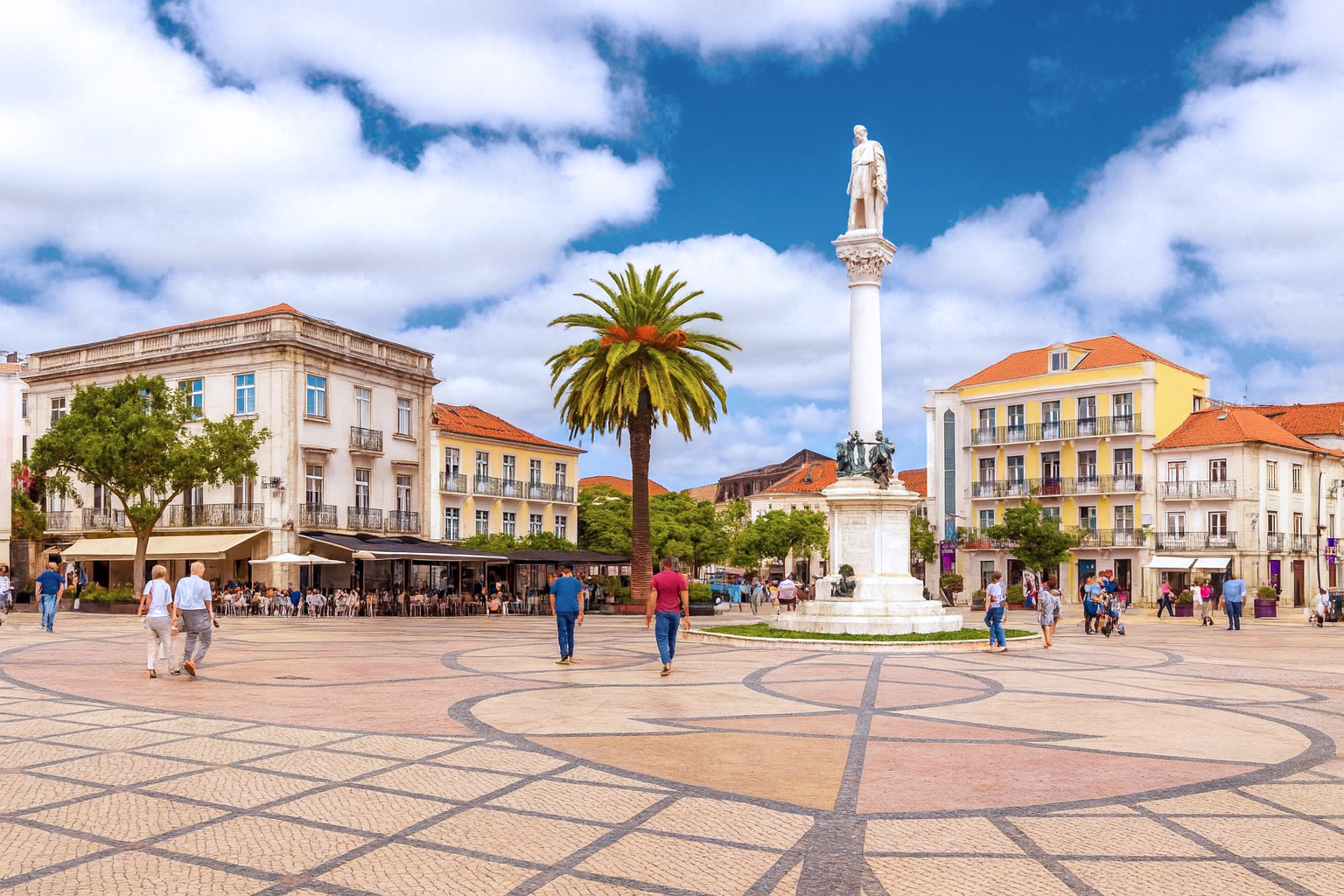 Praça do Bocage in Setúbal mit der Statue des Dichters Bocage, Palmen und bunten Gebäuden bei klarem Sonnenlicht und malerischen weiß-grauen Wolken, während Menschen über den Platz spazieren.