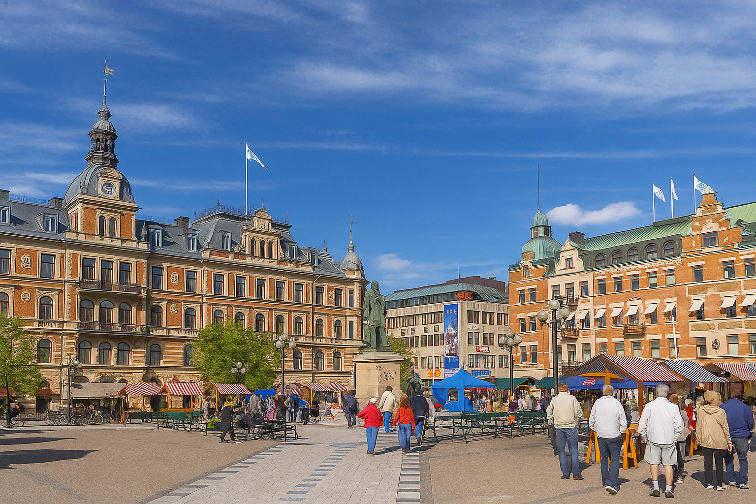 Hauptplatz in Sundsvall mit der historischen Stadthalle und umliegenden Gebäuden aus rotem Backstein, Marktständen und Spaziergängern unter blauem Himmel mit weißen Wolken.