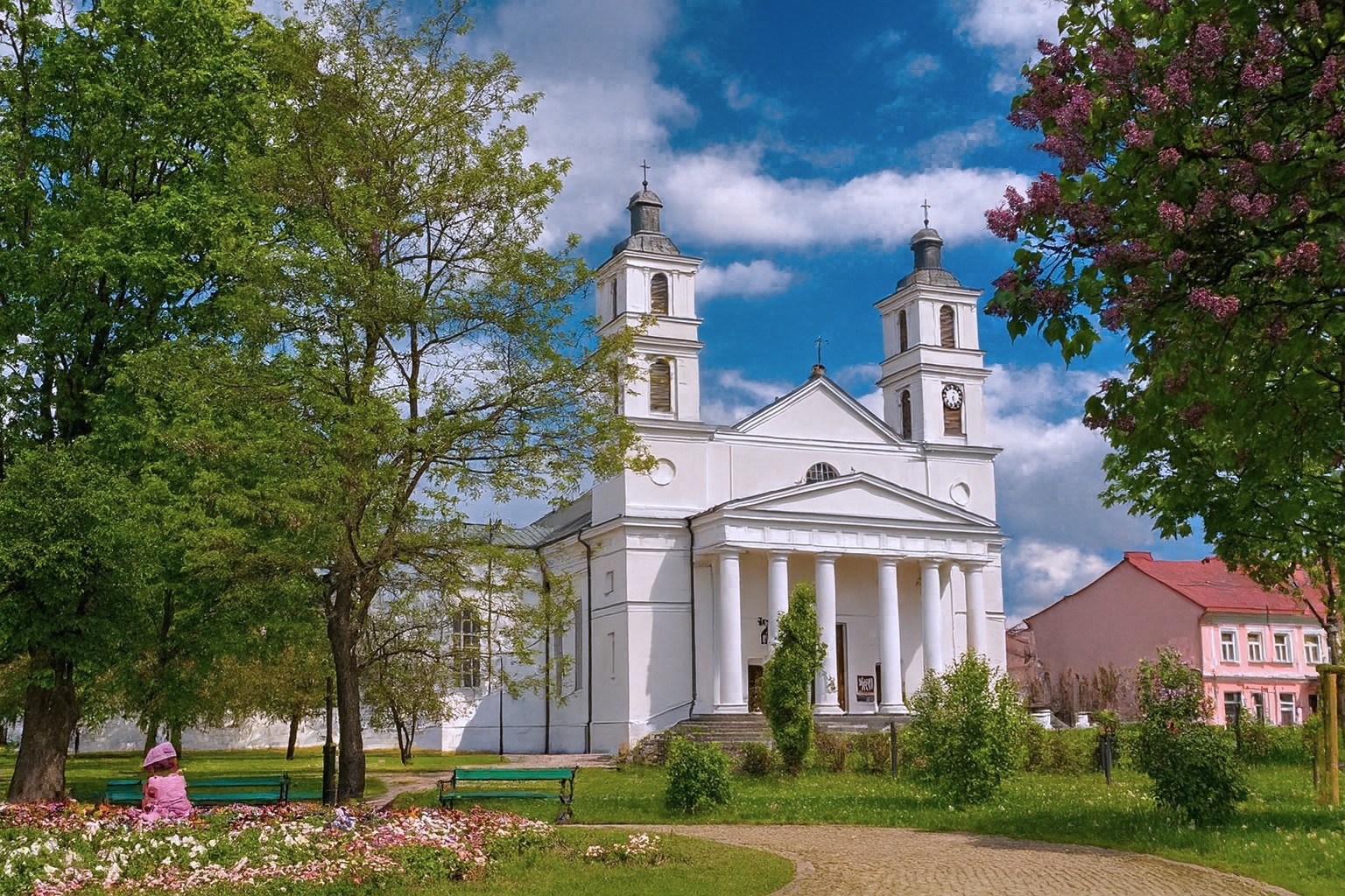Kathedrale in Suwałki in Polen mit zwei Türmen und klassizistischer Fassade bei bestem Sonnenlicht und malerischen Wolken, umgeben von Bäumen und Blumenbeeten.