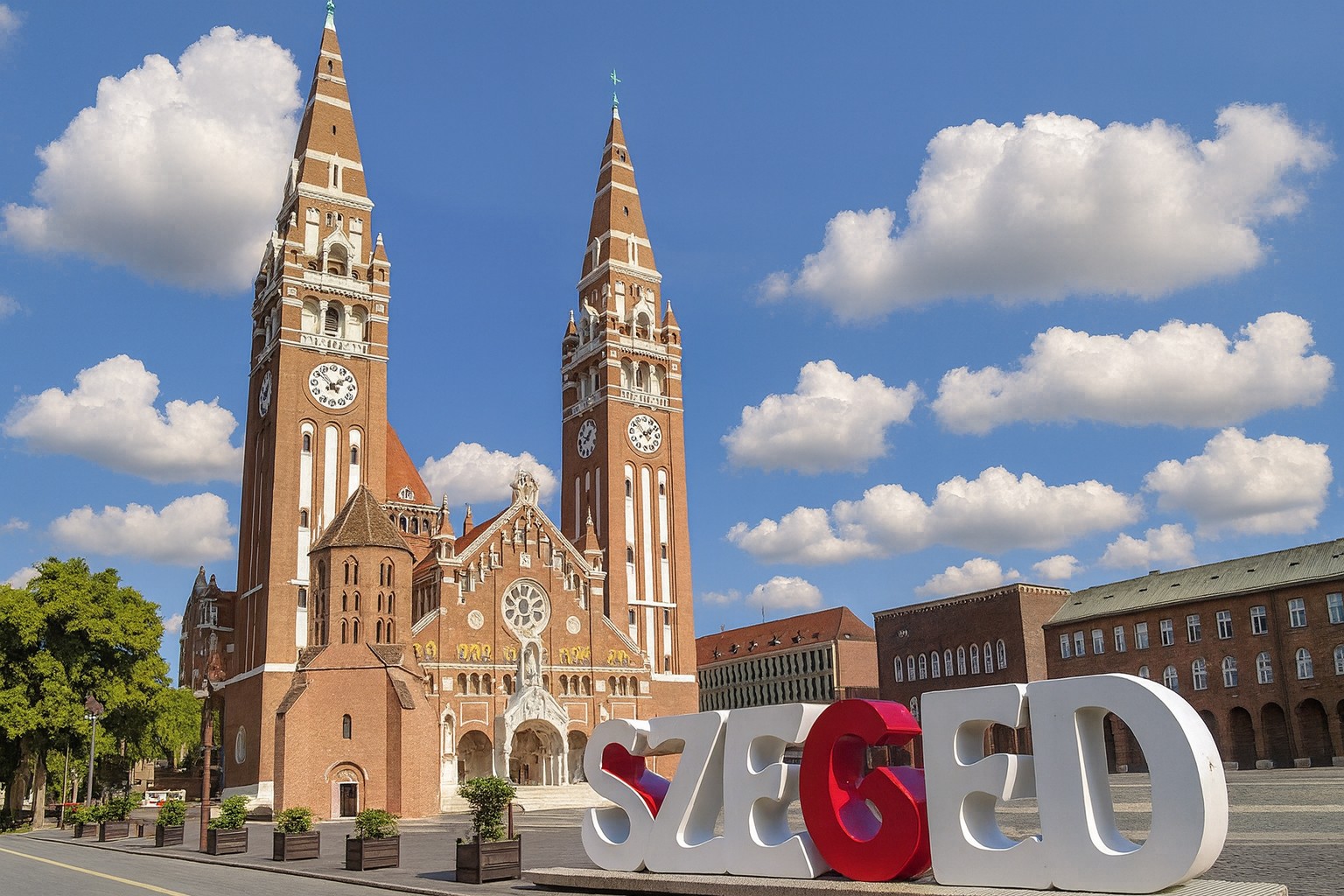 Kathedrale von Szeged in Ungarn mit zwei hohen Türmen und dem Schriftzug „Szeged“ im Vordergrund bei blauem Himmel mit weißen Wolken.