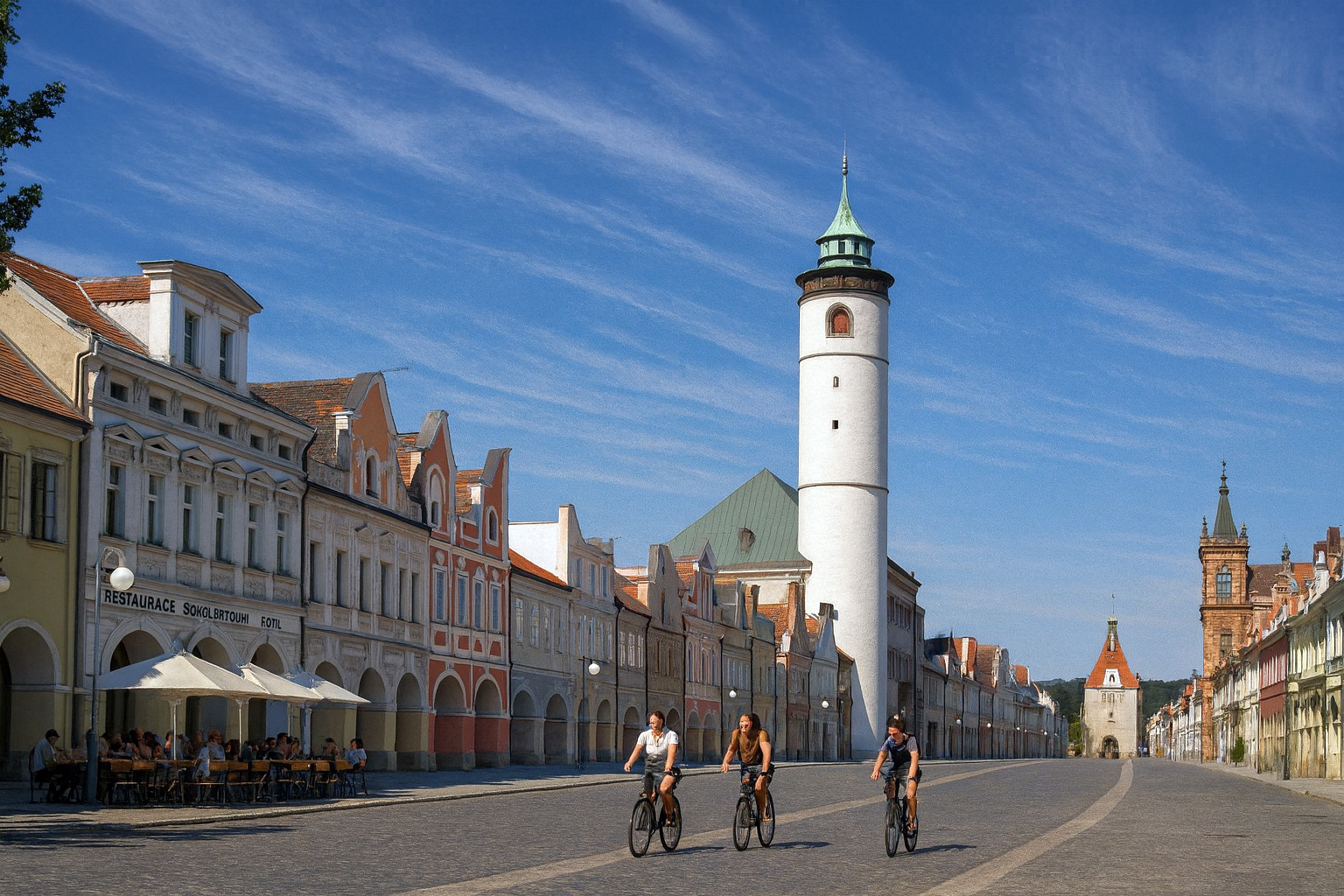 Hauptstraße Náměstí Míru in Taus mit historischem weißen Turm, Straßencafe auf der linken Seite, Spaziergängern und Fahrradfahrern bei strahlender Mittagssonne mit malerischen Wolken.