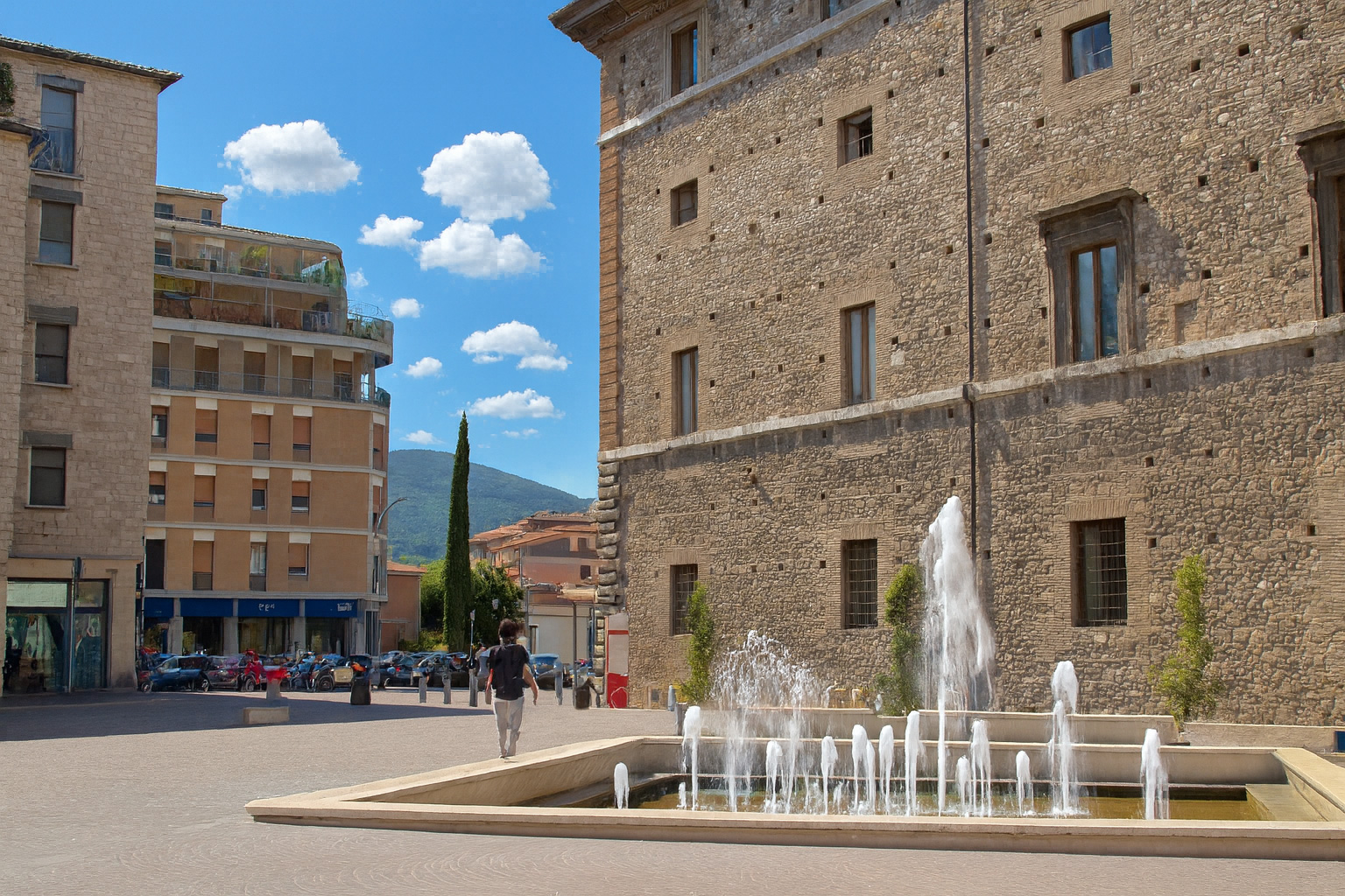 Piazza Europa in Terni mit modernem Brunnen und historischen Gebäuden unter malerischen weiß-grauen Wolken im Sonnenschein.