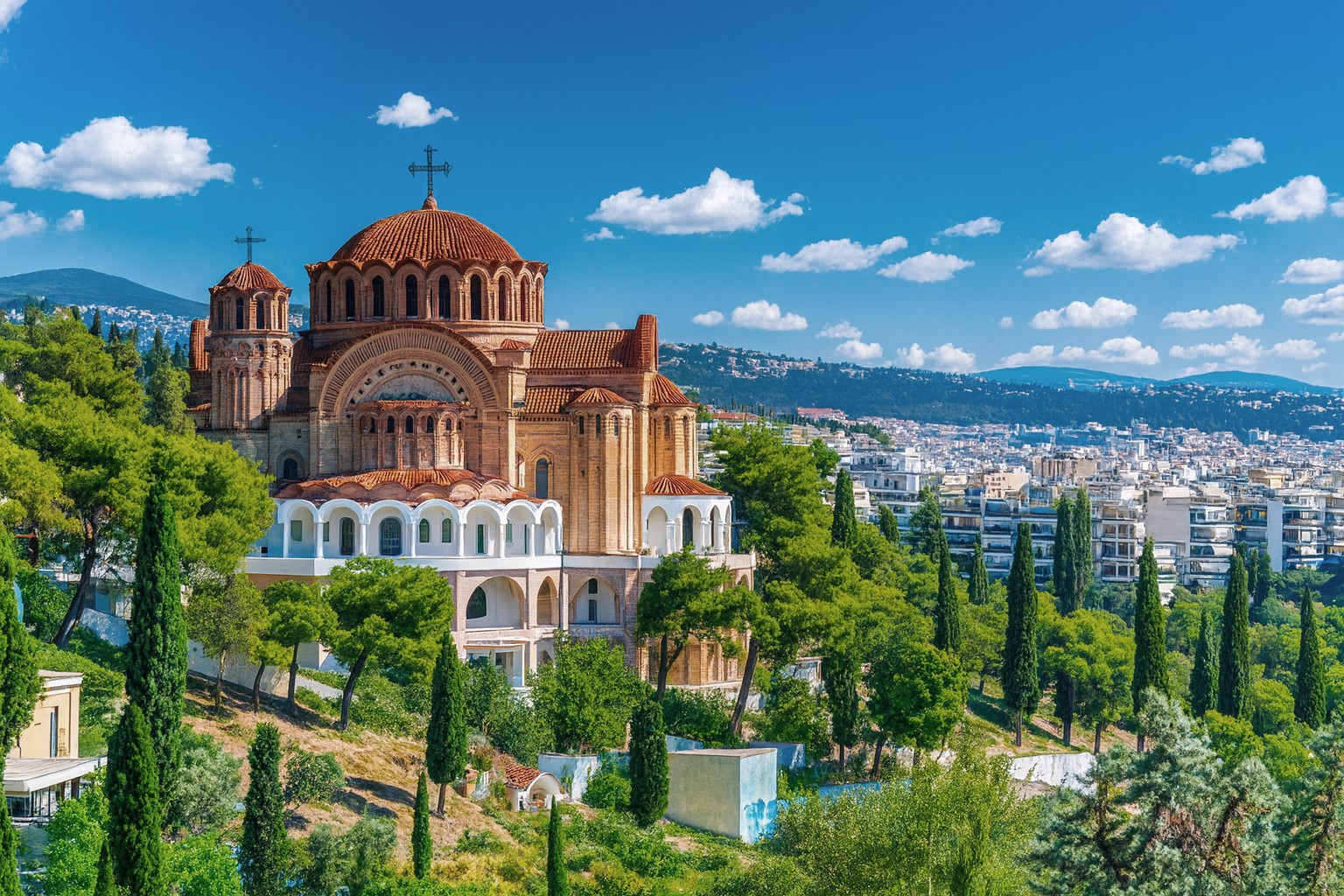Orthodoxe Kathedrale in Thessaloniki mit roten Kuppeldächern, umgeben von Bäumen und Stadtpanorama bei blauem Himmel mit weißen Wolken.