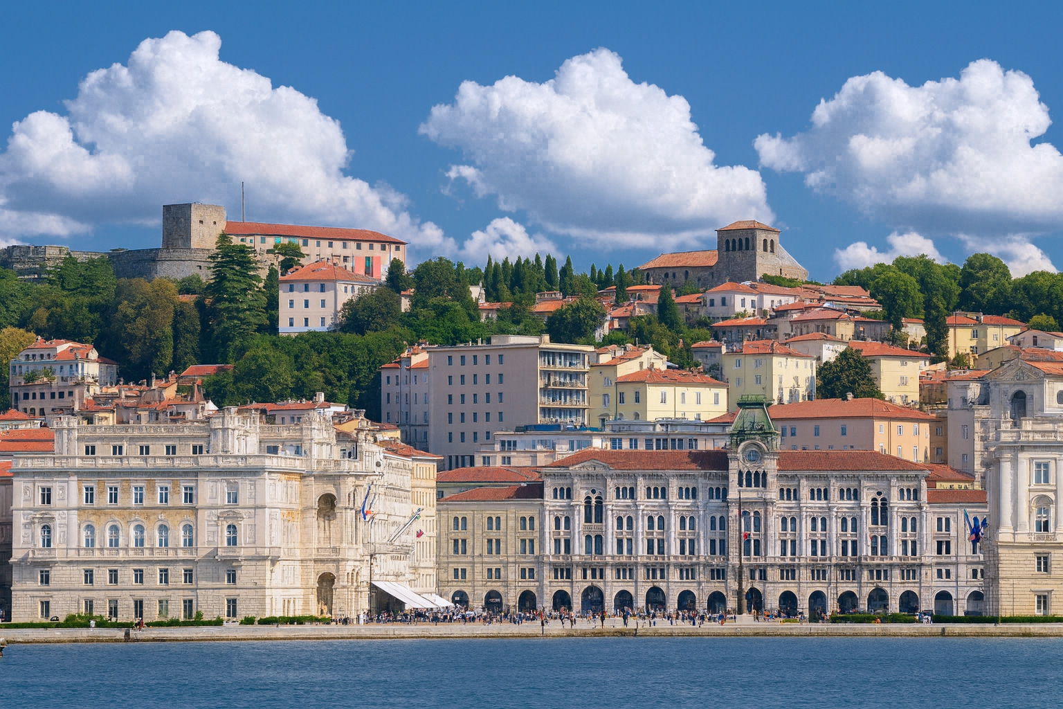 Blick auf die Altstadt von Triest mit historischen Palästen an der Uferpromenade und dem Castello di San Giusto im Hintergrund bei blauem Himmel mit weißen Wolken.