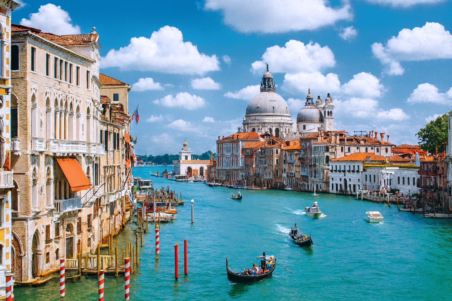 Blick auf den Canal Grande in Venedig mit der Basilika Santa Maria della Salute, Gondeln und Booten bei blauem Himmel mit weißen Wolken.