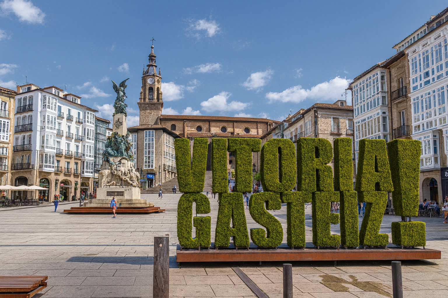 Andre Maria Zuriaren Plaza in Vitoria-Gasteiz mit der grünen VITORIA-GASTEIZ-Schriftinstallation, dem Denkmal im Hintergrund und Spaziergängern bei sonnigem Himmel mit weißen Wolken.