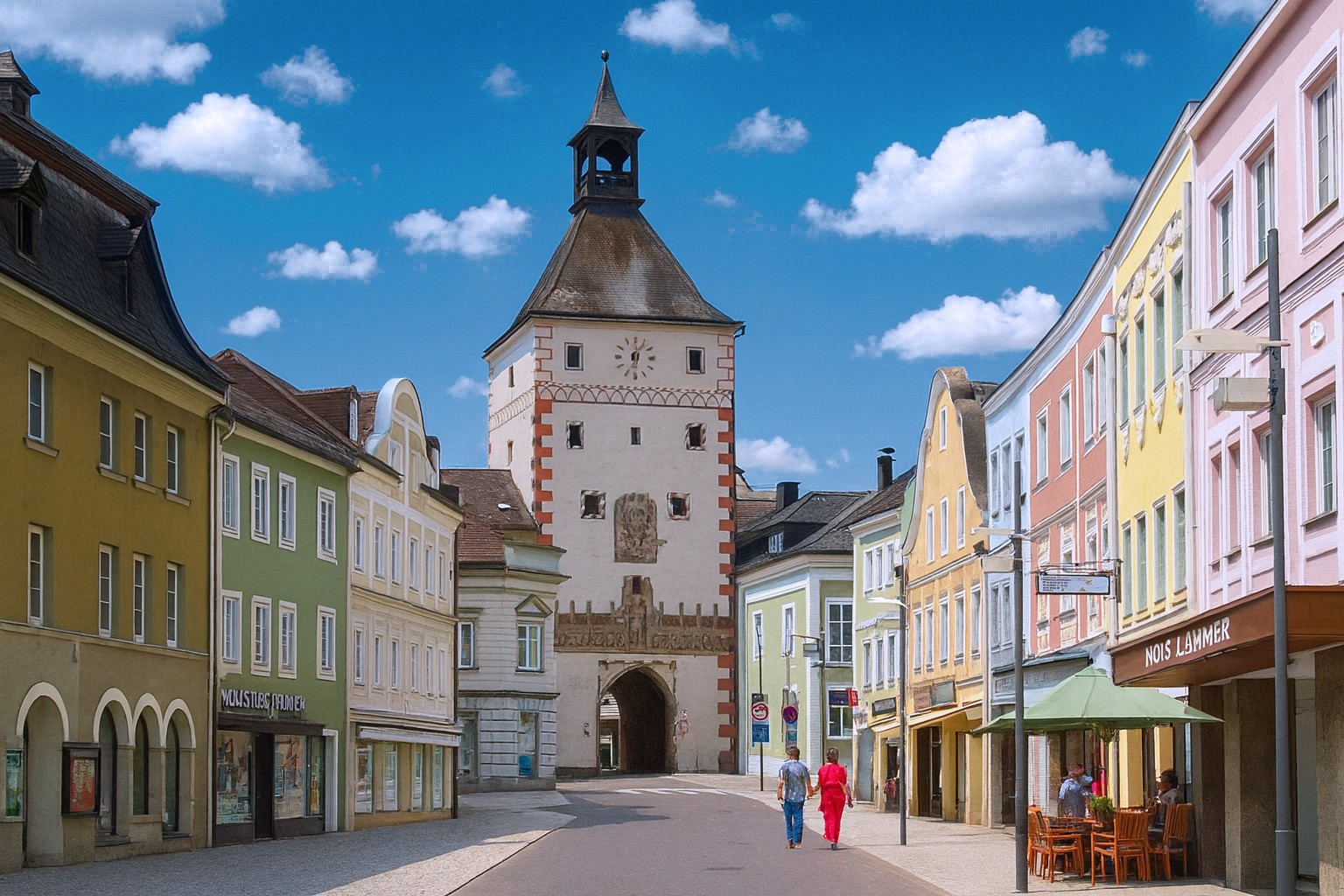 Altstadt von Vöcklabruck mit dem historischen Stadtturm im Hintergrund, bunten Fassaden, Straßencafe und Spaziergängern.