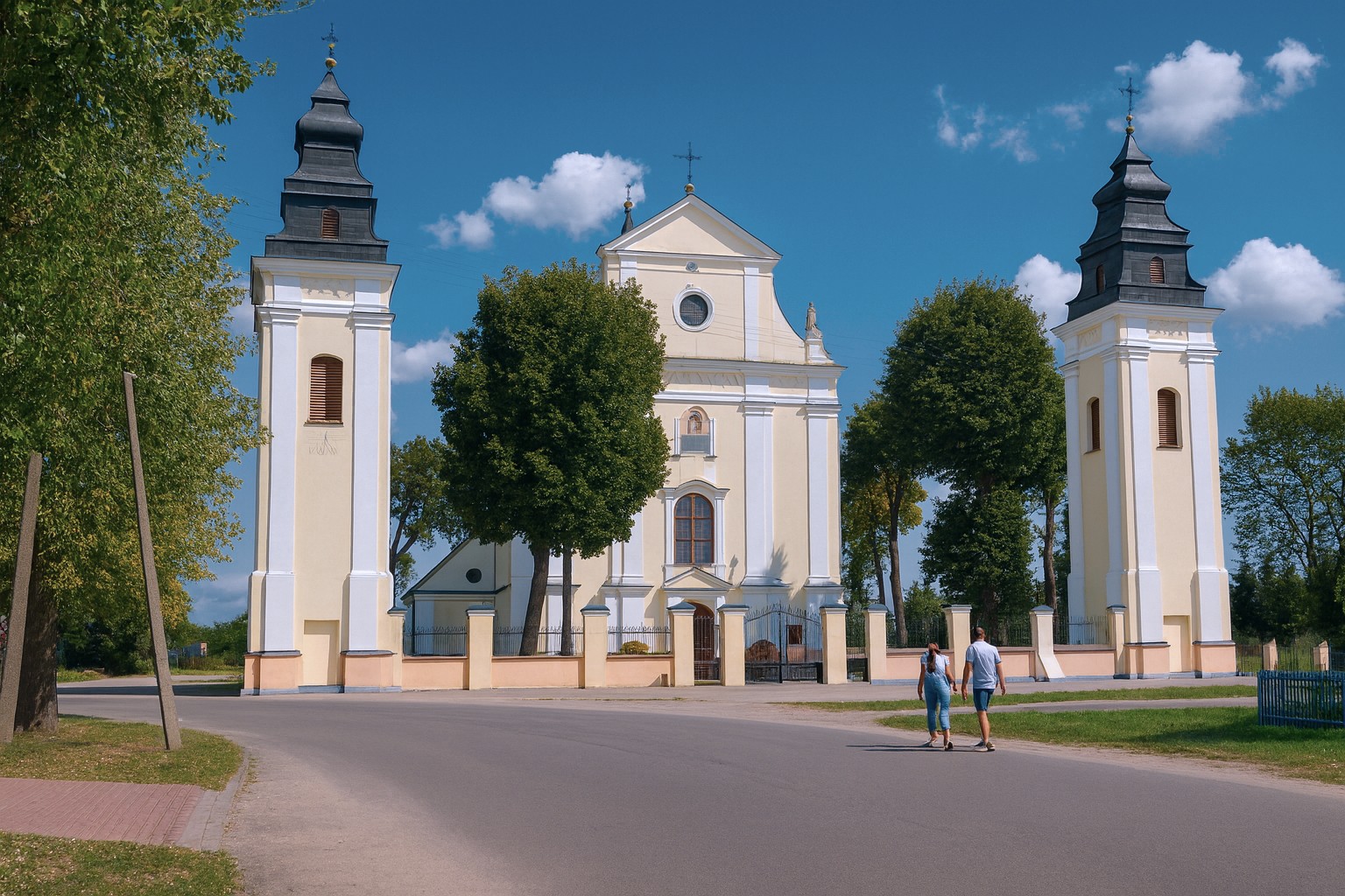 Kirche in Wyszków in Polen mit zwei Türmen und klassizistischer Fassade bei bestem Sonnenschein und malerischen Wolken, zwei Personen gehen auf das Kirchenportal zu.