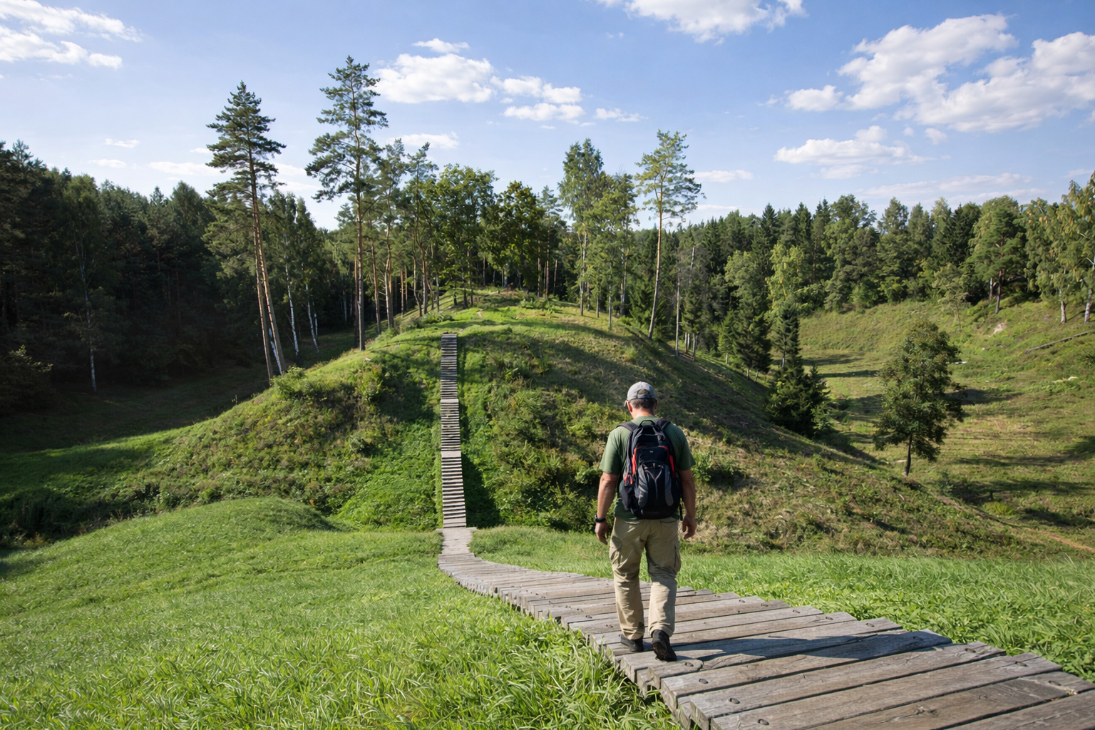 Holzsteg in einer offenen Hügellandschaft im Žuvintės kraštovaizdžio draustinis in Litauen mit steilem Treppenabstieg, Wiesenhängen, einzelnen Kiefern und dichtem Waldrand unter blauem Himmel mit lockeren Wolken, ein Wanderer geht den Holzweg hinunter