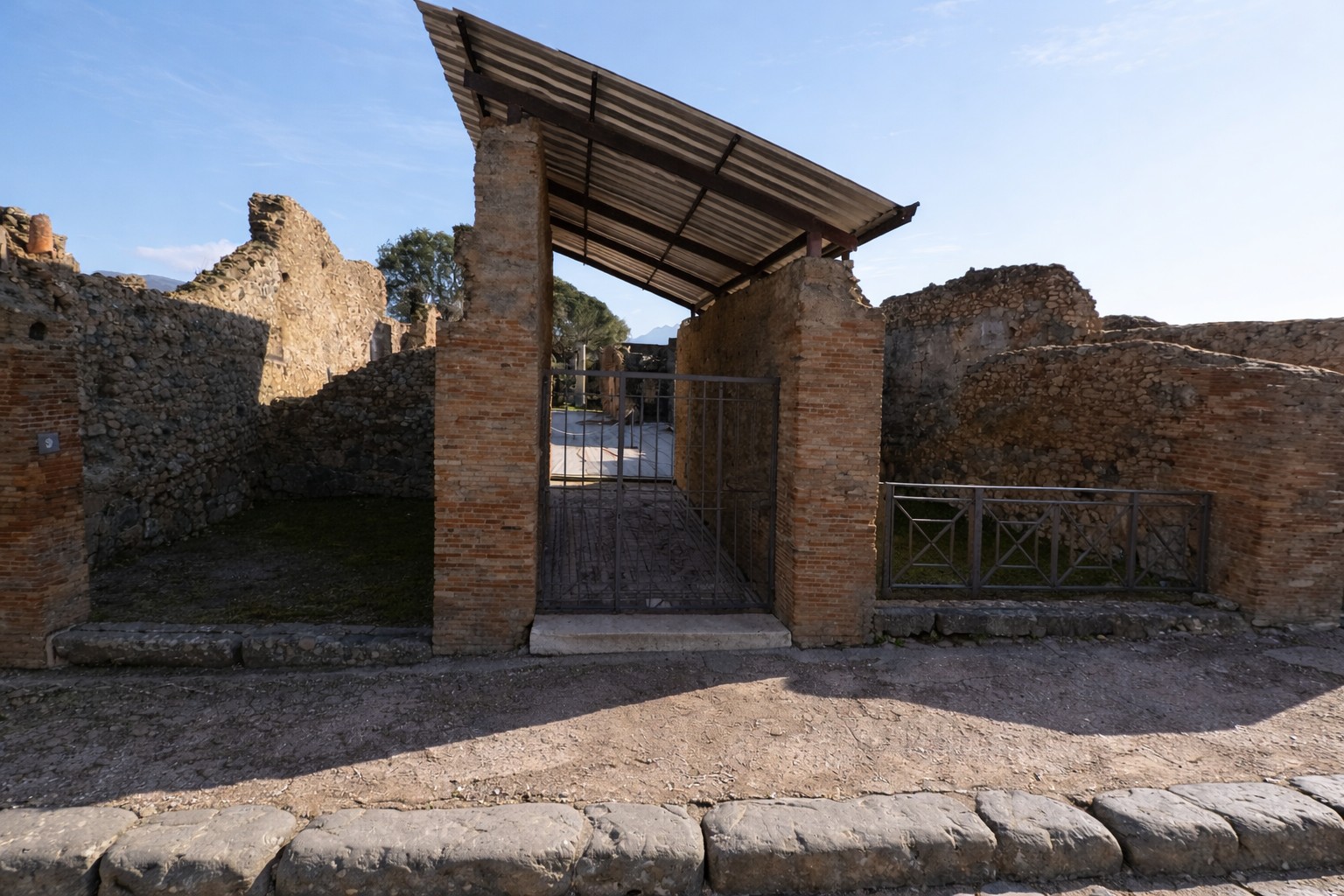 Eingang der Casa del Jabalí in Pompei mit antiken Ziegelpfeilern, überdachtem Zugang und erhaltenen Mauerresten entlang der gepflasterten Straße