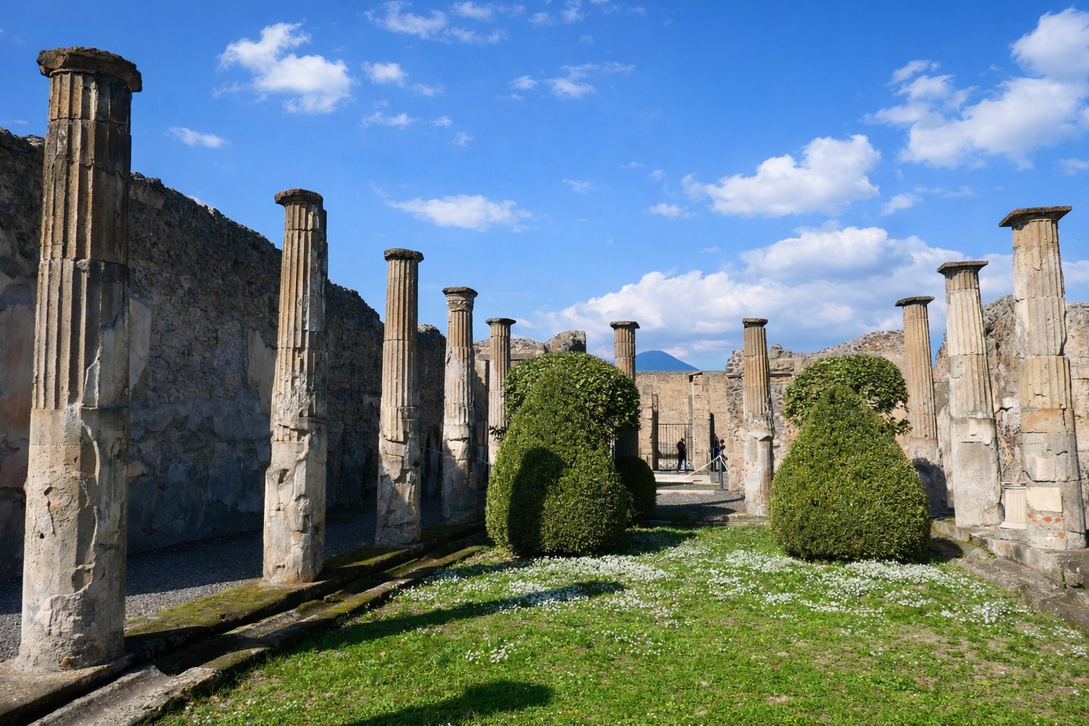 Peristyl der Casa dei Cornelii in Pompei mit antiken Säulenreihen, gepflegten Buchsbäumen im Innenhof und Blick auf die umliegenden Ruinen unter leicht bewölktem Himmel