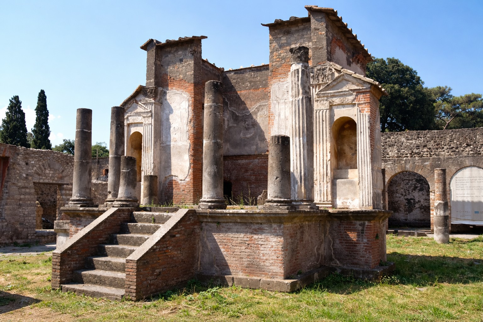 Tempio di Iside in Pompei mit erhobenem Podium, Treppenaufgang und teilweise erhaltenen Säulen, Blick auf die reich gegliederten Tempelwände und Nischen unter klarem Himmel