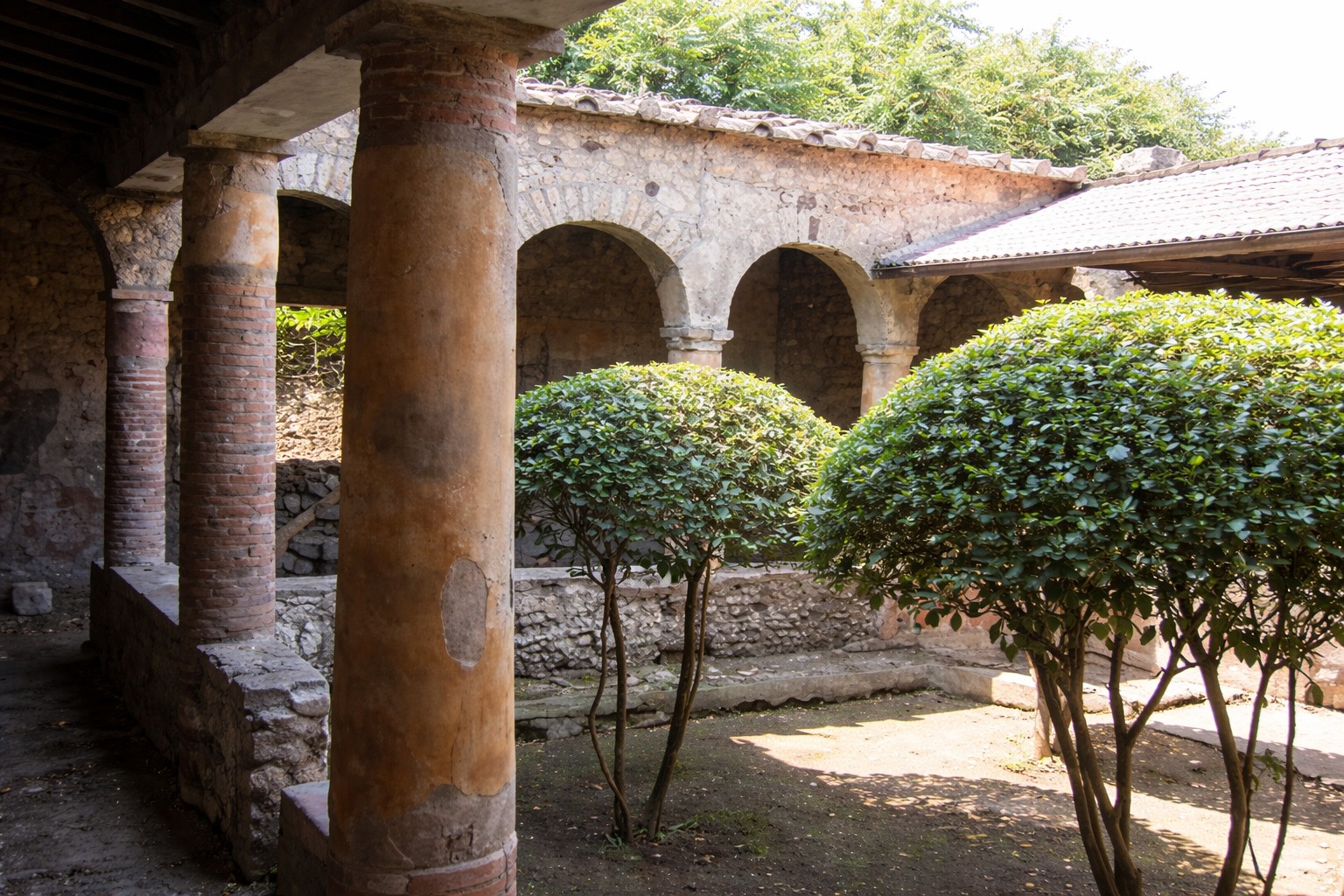 Innenhof der Casa degli Archi in Pompei mit überdachtem Säulengang, gemauerten Bögen und dicht geschnittenen Sträuchern, Blick durch den schattigen Portikus auf die antike Gartenanlage