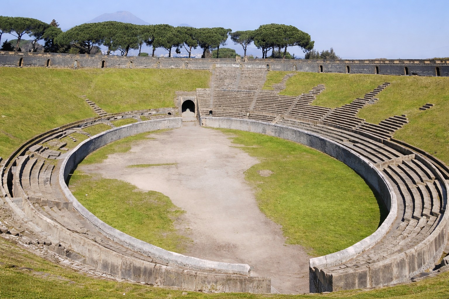Anfiteatro di Pompei mit ovaler Arena, umlaufenden steinernen Zuschauerrängen und Blick auf die erhaltenen Stufenanlagen vor Pinien und dem Vesuv im Hintergrund